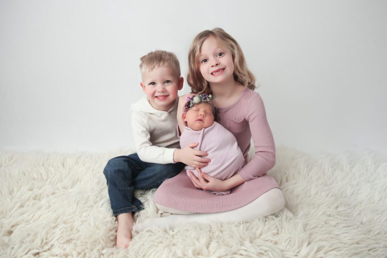 Brother and sister hold their newborn sister on a fuzzy beige rug during a baby photography session.
